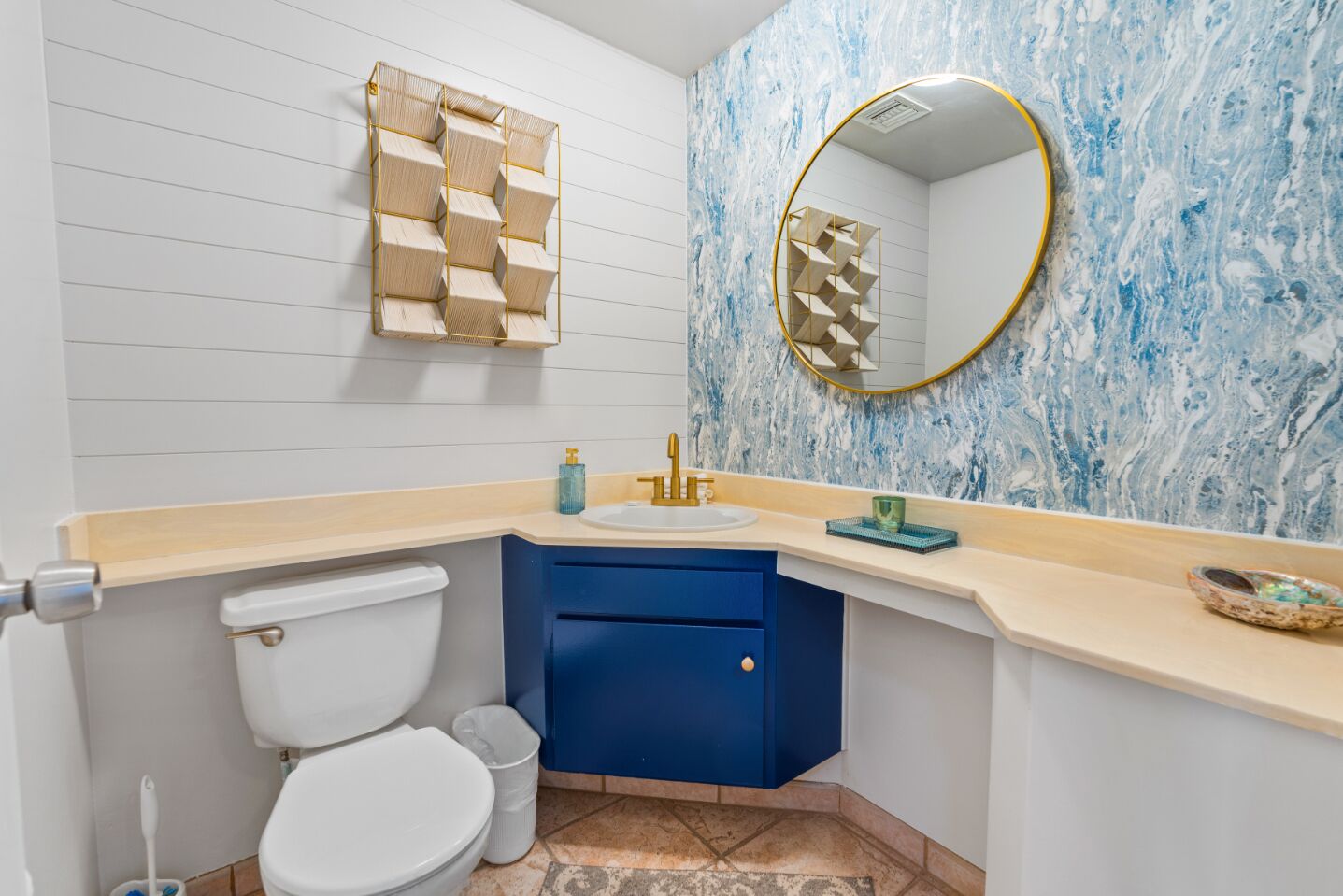 A bathroom with a white toilet, a blue cabinet, a sink with a gold faucet, and a marble wall with a gold-framed mirror.
