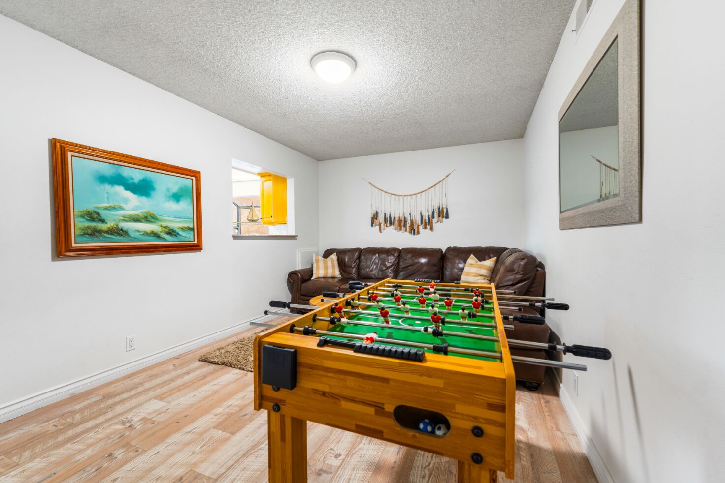 A room with a foosball table, a brown leather sofa, a framed painting, and a window with a view.