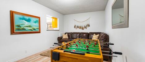 A room with a foosball table, a brown leather sofa, a framed painting, and a window with a view.