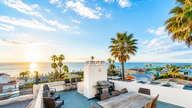 An outdoor patio with a view of the ocean, a grill, wicker furniture, and a white wall with a clock.