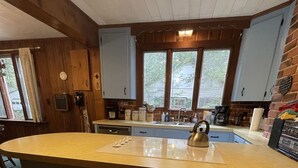 Una cuina amb una illa central, dues cadires de fusta, un rentapeus i una finestra amb vista a vegetació.