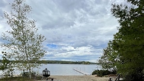 Taula de picnic de fusta amb una graella en una platja sorrenca, envoltada d'arbres i un llac.