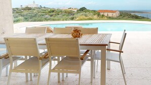 Un espace extérieur aménagé, abrité et doté de mobilier en bois, offrant une vue sur la piscine et s’ouvrant sur un ciel dégagé.
