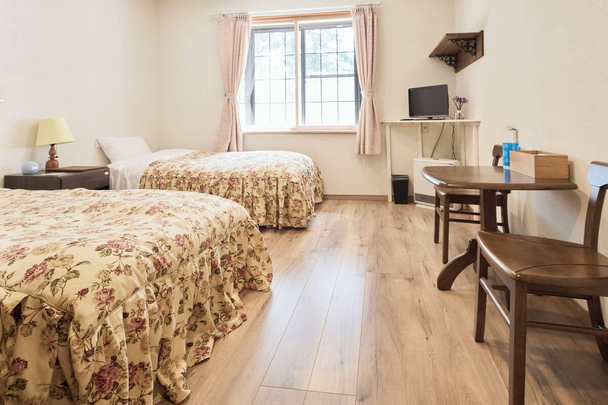 A room with two beds, a desk, a chair, a TV, and a window with curtains.