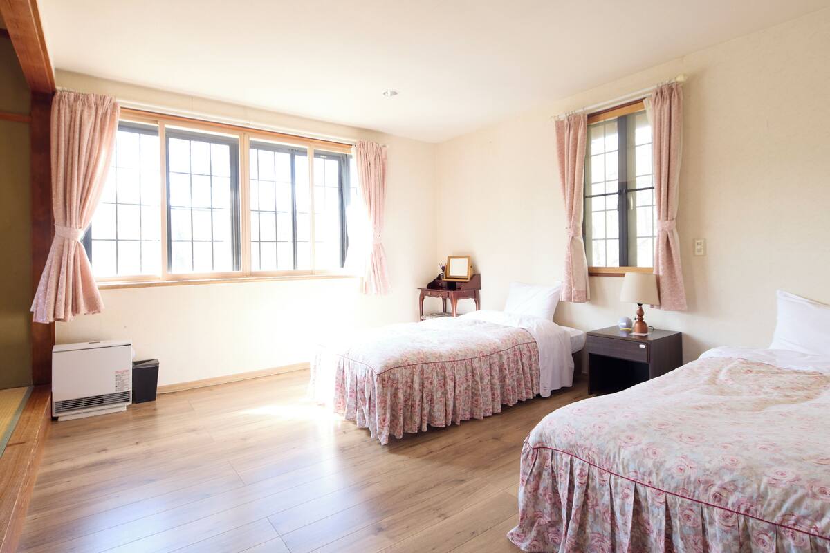 A room with two beds, large windows, a heater, and a small table with a lamp.