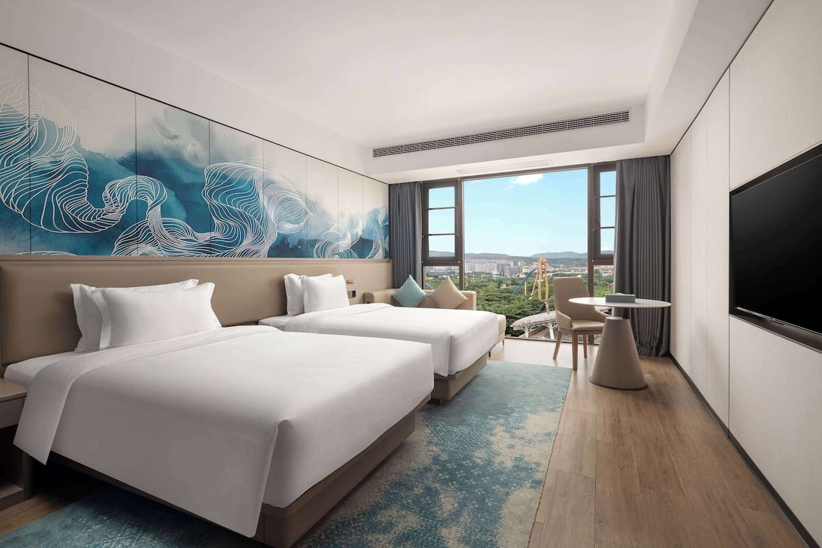A hotel room with two beds, a large window with a city view, a flat-screen TV, and a small table.