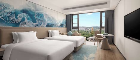 A hotel room with two beds, a large window with a city view, a flat-screen TV, and a small table.