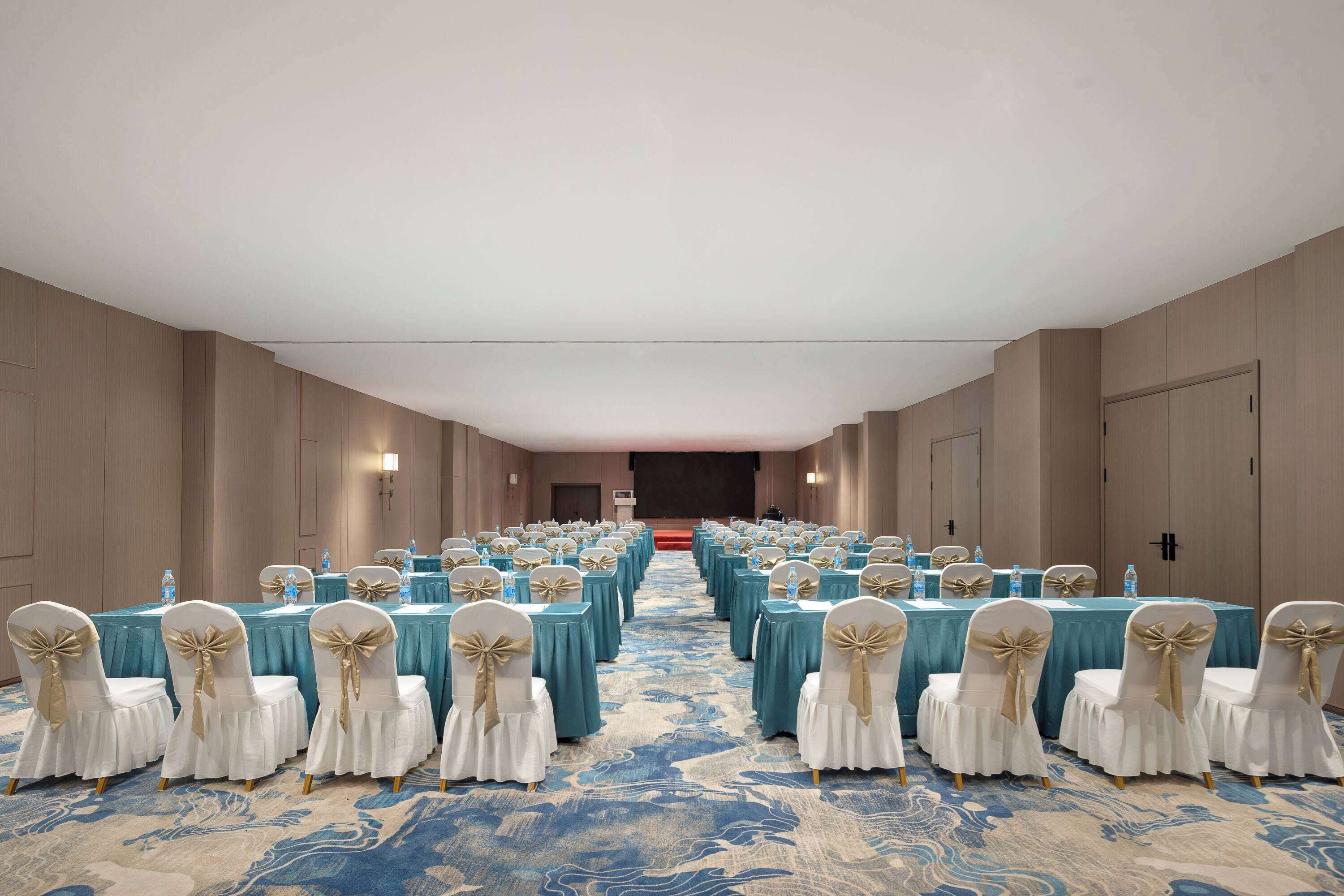 A conference room with rows of tables and chairs, a carpeted floor, and a stage at the front.