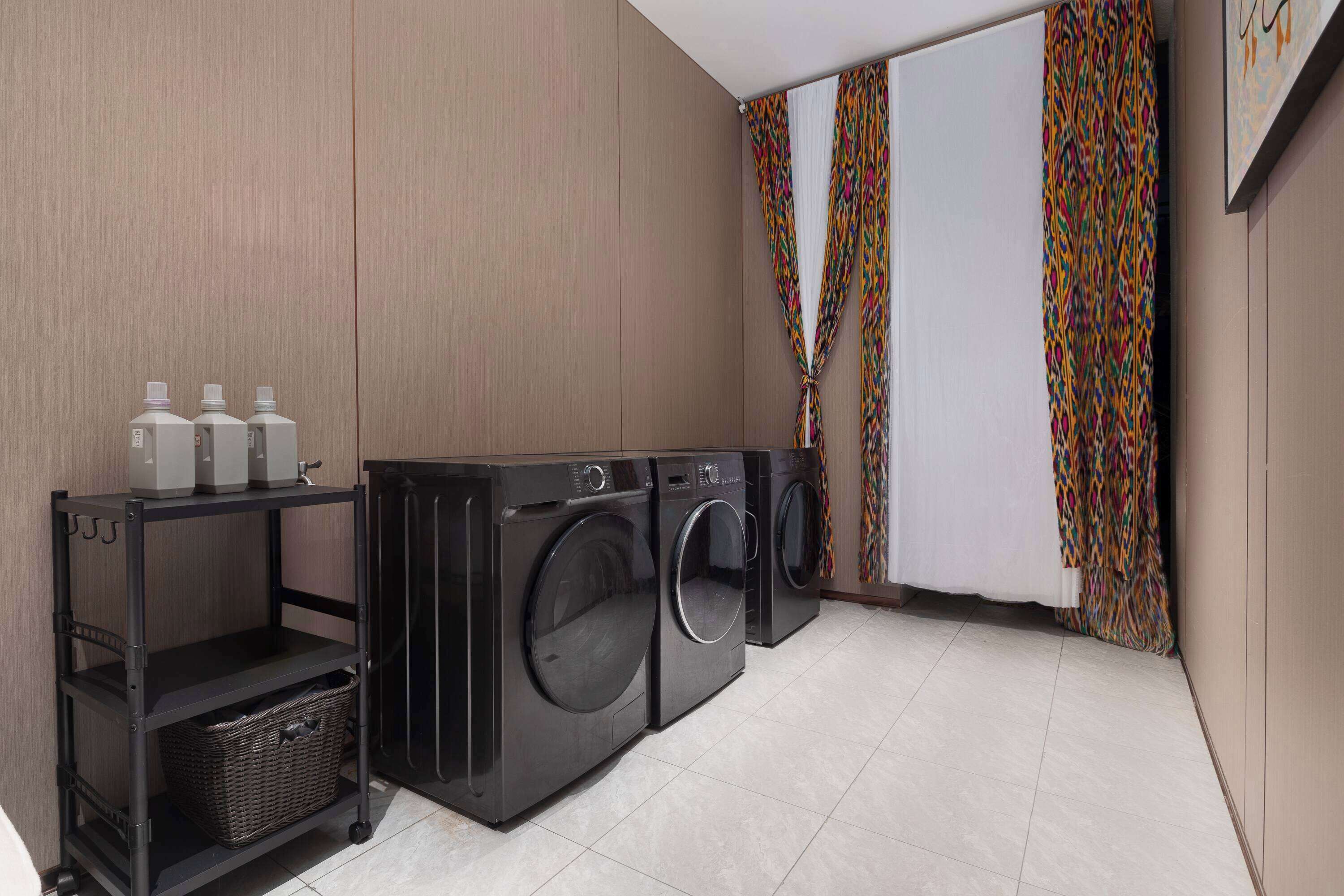 A laundry area with two front-loading washing machines, a black metal shelf with bottles, and a wicker basket.