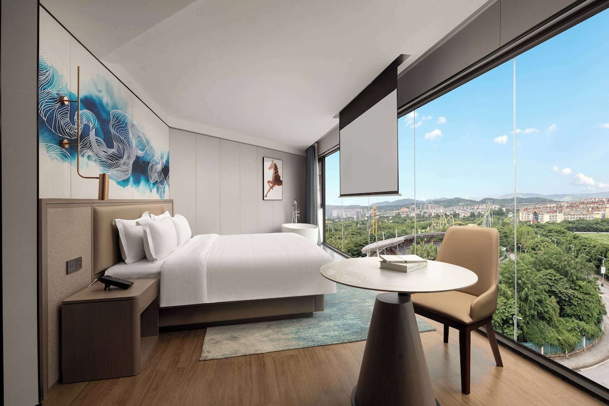 A modern hotel room with a large bed, a bedside table, a chair, a round table, and a large window offering a city view.