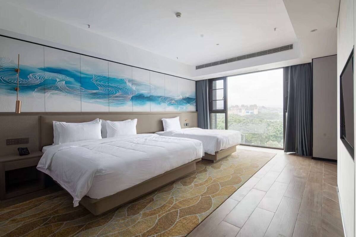 A modern hotel room with two beds, a large abstract painting on the wall, a large window with a city view, and a patterned rug.