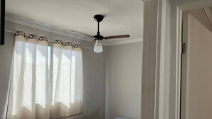 Ein Schlafzimmer mit einem Einzelbett, einem Deckenventilator und durchsichtigen Vorhängen an einem Fenster.