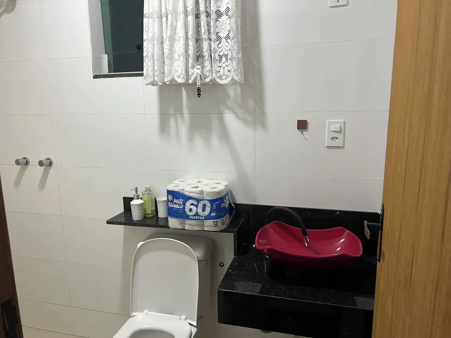 A small bathroom with a toilet, a sink with a red basin, and a shelf with toilet paper and toiletries.