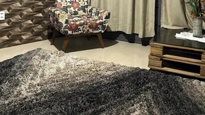 Une pièce avec un mur orné de motifs, un fauteuil à fleurs, un grand tapis et une petite table en bois sur laquelle est posé un vase.
