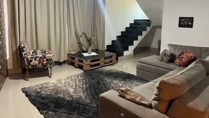 Un salon moderne comprenant un canapé, une table basse, un fauteuil, un escalier et une grande fenêtre avec des rideaux.