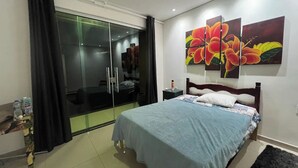 Une chambre à coucher comprenant un lit, une table de chevet, une porte vitrée donnant sur une salle de bain et un mur orné de trois tableaux représentant des motifs floraux.
