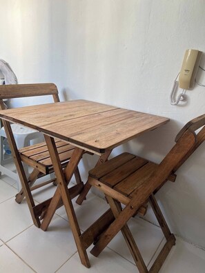 Simple table and chairs sit against a plain wall, accompanied by a mounted telephone and a tiled floor.