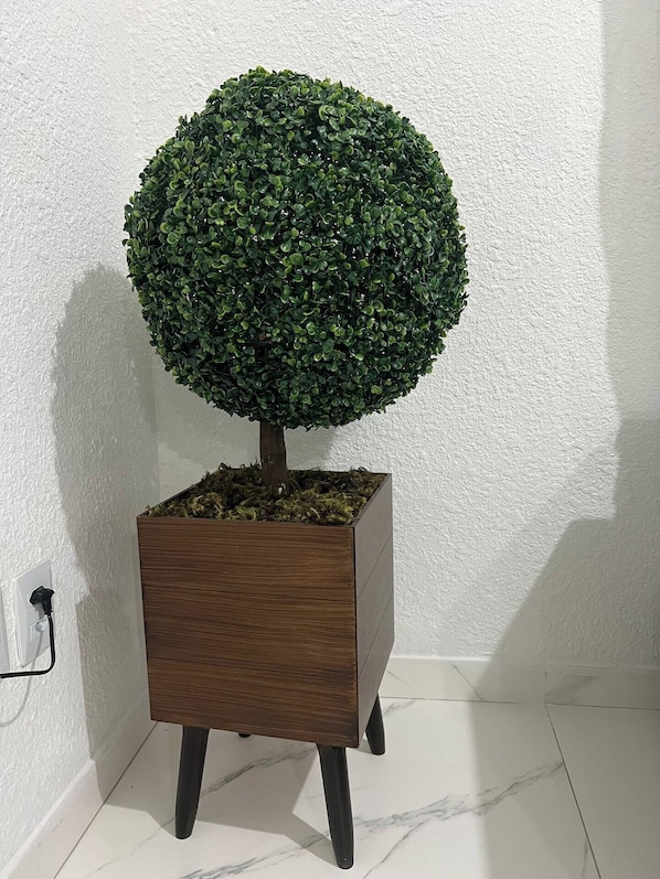 Un árbol en maceta con jardinera de madera sobre un suelo de baldosas frente a una pared blanca.
