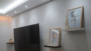 A modern living room with a flat-screen TV mounted on the wall, surrounded by minimalist decor including a framed picture, a small sculpture, and a bonsai tree on floating shelves. - 1 Near Confins Airport, Cid Galo, 15 min BH, Cid Adm.  (Centro Vespasiano)