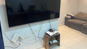 Un televisor de pantalla plana montado en una pared, una estantería de madera con dispositivos electrónicos, y un sofá gris en una sala de estar.