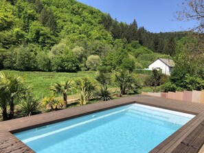 A rectangular swimming pool with a wooden deck, surrounded by lush greenery and a hillside.