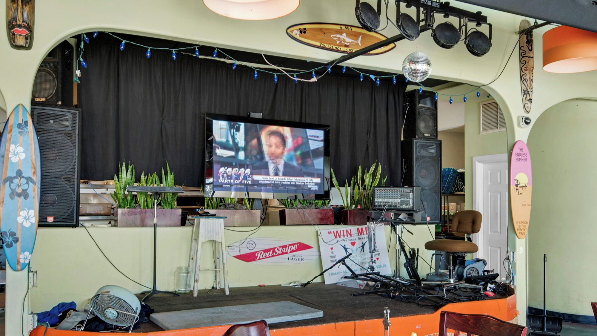 Een podium met een televisie waarop een sportevenement wordt vertoond, omringd door luidsprekers, een drumstel en sierplanten.