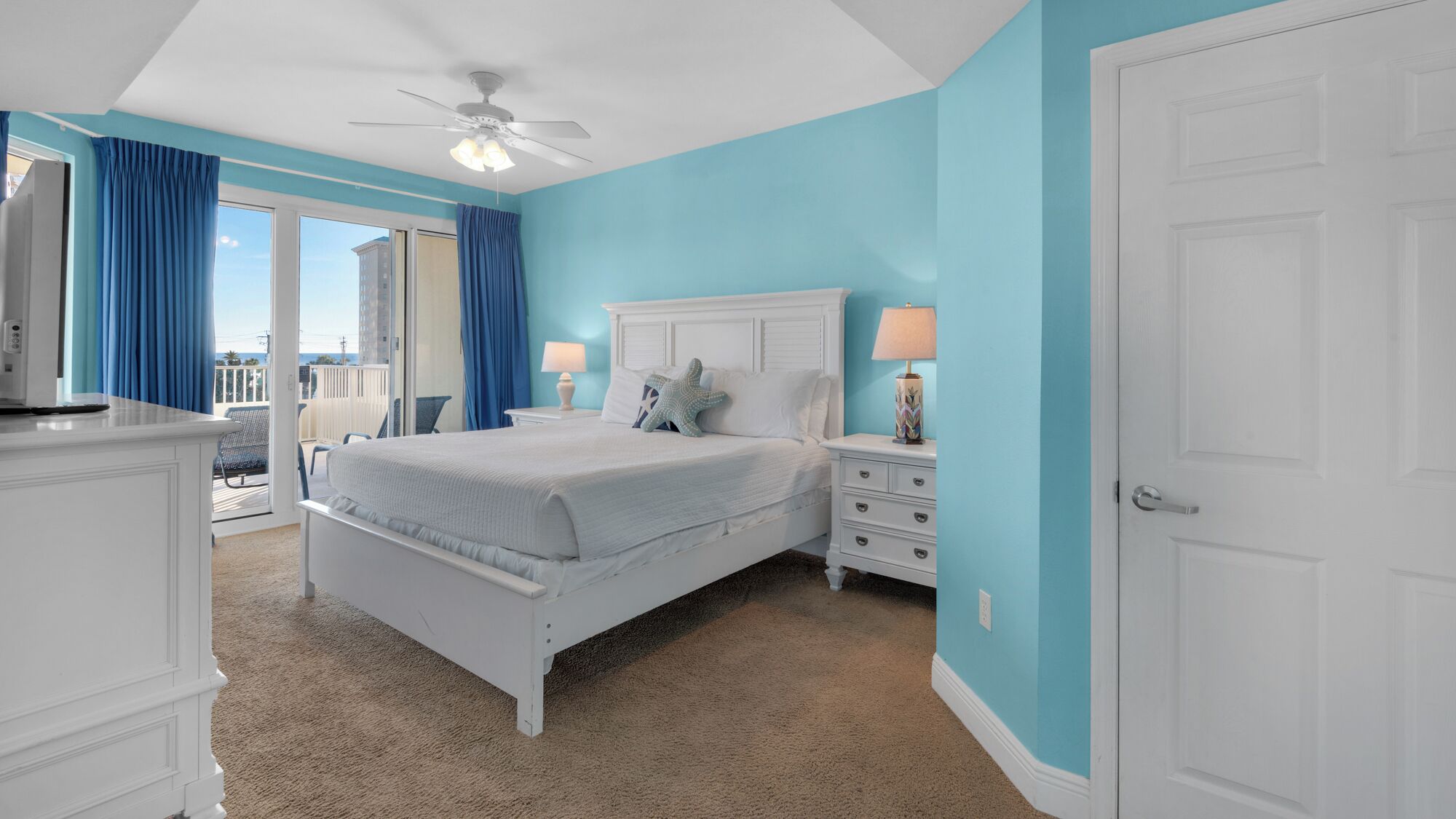 Une chambre à coucher comprenant un lit, des tables de chevet, une commode, un ventilateur de plafond et une vue sur l’extérieur.