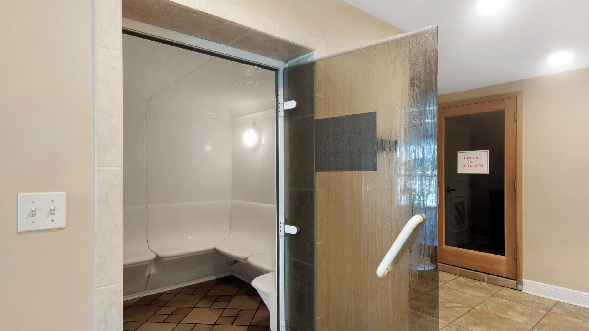 Une salle de bain avec une douche à parois de verre, des toilettes et un lavabo. Un panneau sur la porte indique qu'il est obligatoire de porter un maillot de bain.