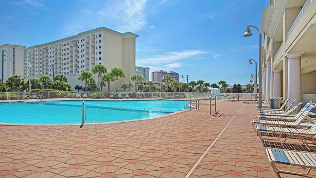 Una piscina con sillas de descanso, un camino empedrado y un edificio de varios pisos al fondo.