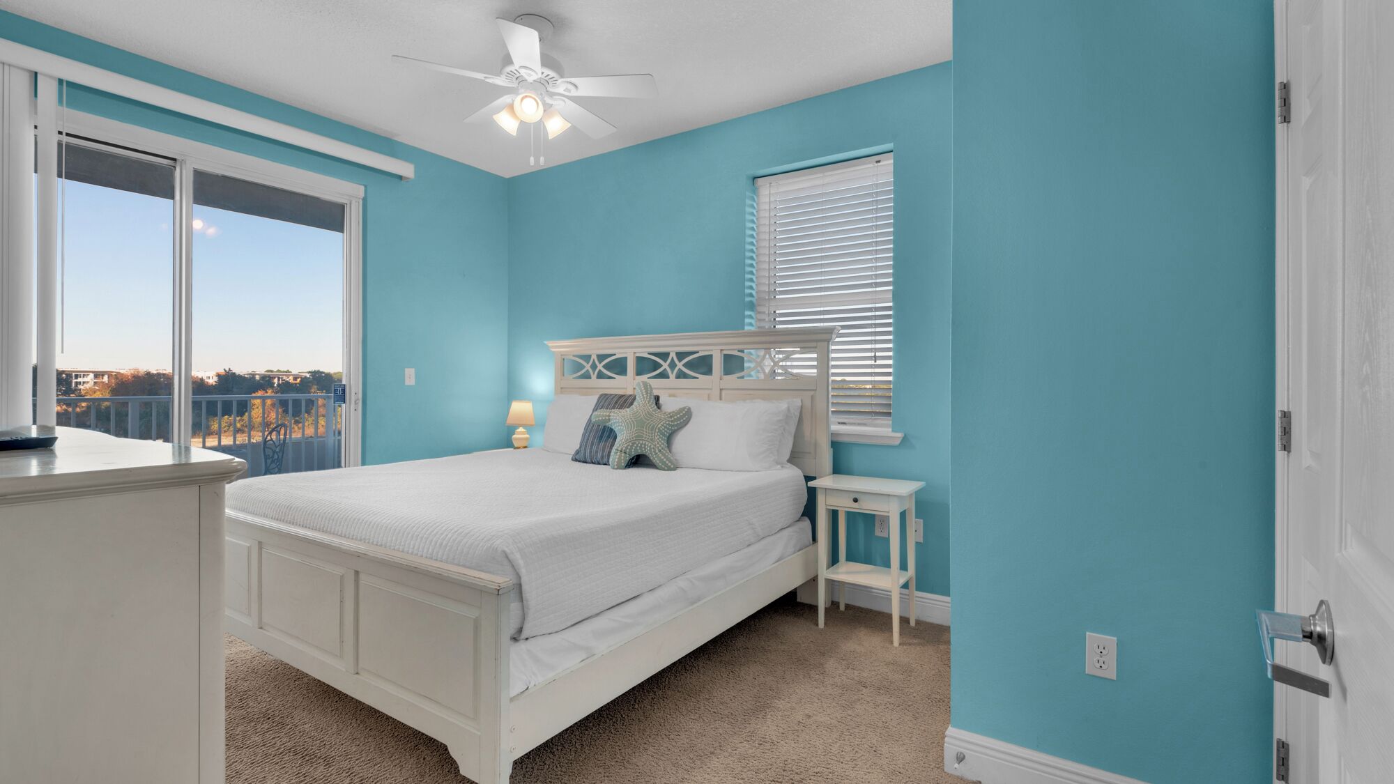 A bedroom with a bed, a nightstand, a ceiling fan, a window with blinds, and a view of the outside.