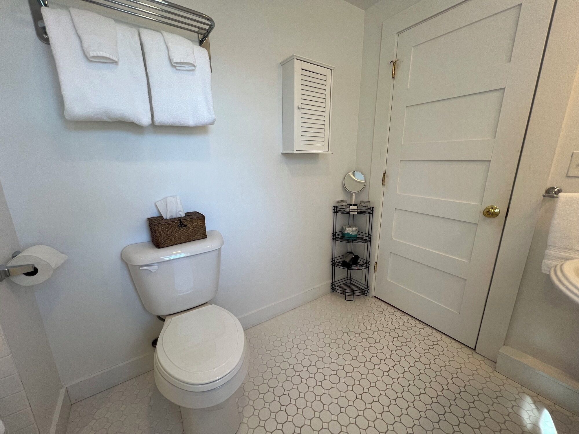 A bathroom with a toilet, a tissue box, a towel rack with two towels, a toilet paper holder, a small shelf with a mirror and toiletries, and a door with a handle.