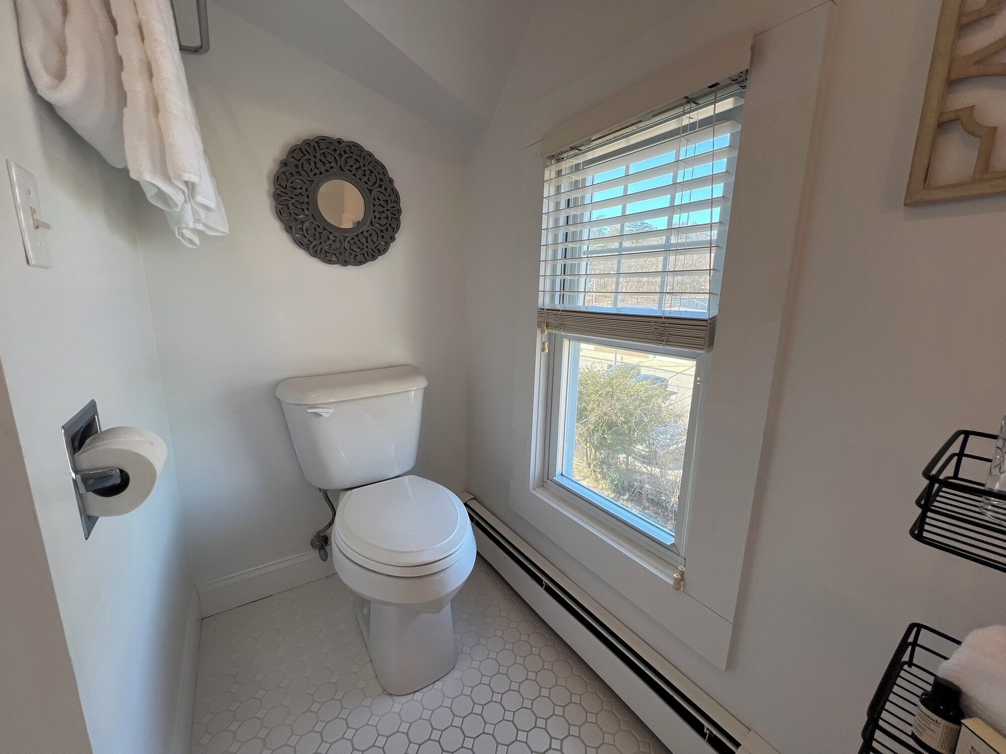 A bathroom with a toilet, a window with blinds, a towel rack, and a decorative wall piece.