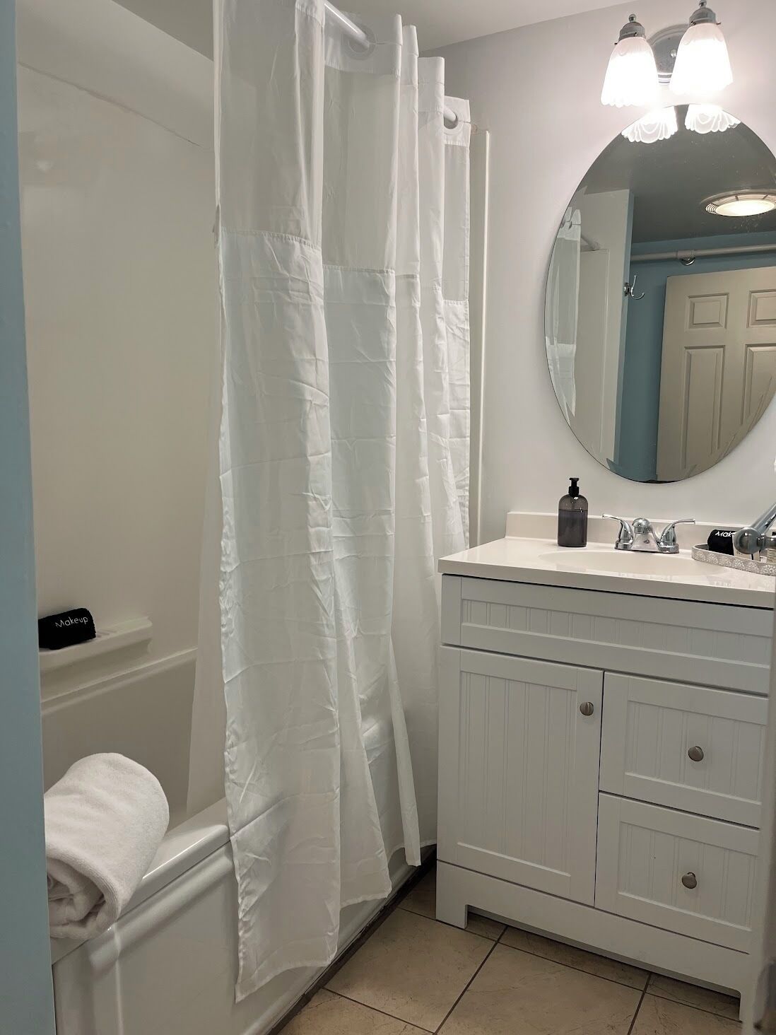 Un bagno con tenda da doccia bianca, uno specchio rotondo, un mobile lavabo bianco con cassetti e un asciugamano sul pavimento.