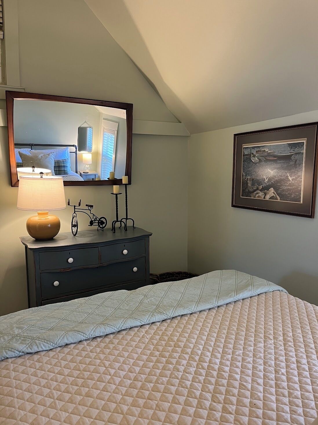 A hotel room with a bed, a dresser with a lamp, a mirror, and a framed picture on the wall.