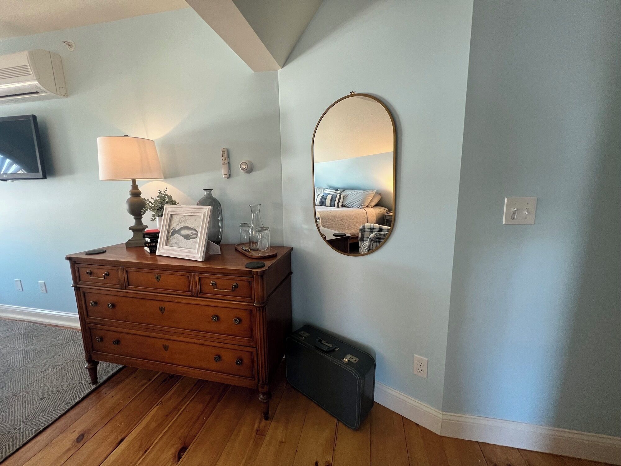 Una habitación con tocador de madera, espejo, lámpara y maleta.