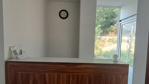 A reception area with a wooden counter, a clock on the wall, and a window offering a view of greenery outside. - JAMUNAS B&B (Shimla)