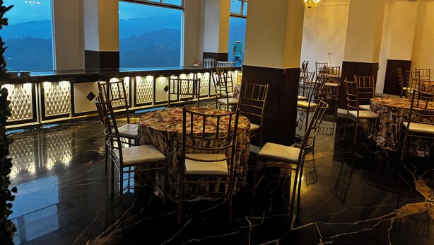 A restaurant with round tables and chairs, large windows with a mountain view, and chandeliers. - JAMUNAS B&B (Shimla)
