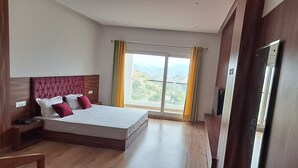 A bedroom with a large bed, a wooden headboard, a nightstand, and a view of the outdoors through a window. - JAMUNAS B&B (Shimla)