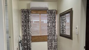 Un pasillo con suelo de madera, una ventana con cortinas estampadas, un equipo de aire acondicionado y un cuadro en la pared.