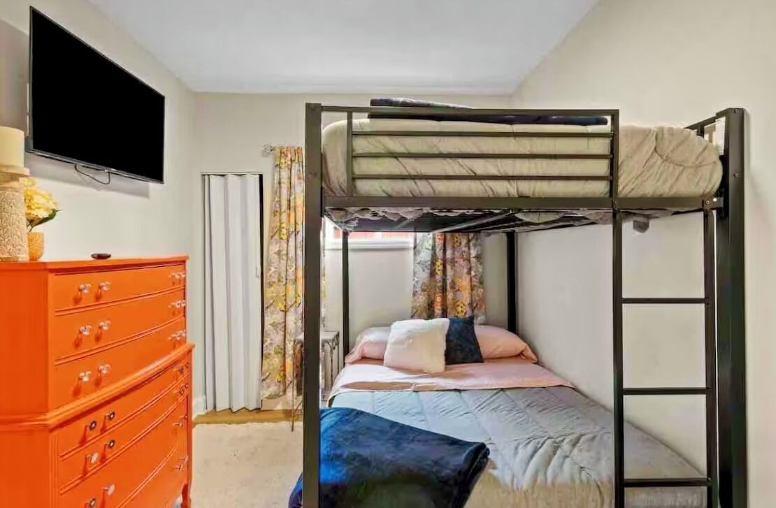 A bunk bed with a TV above it, a dresser, and a window with curtains.