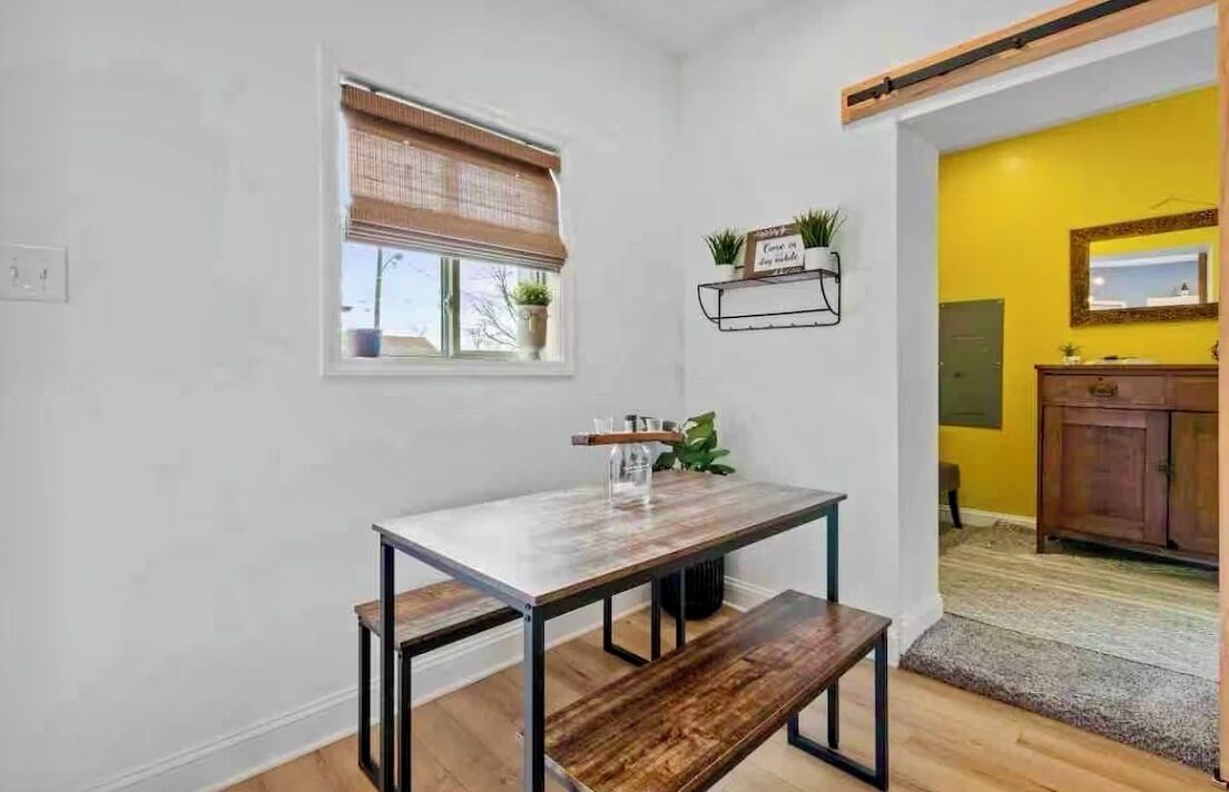 A dining area with a wooden table and bench, a window with blinds, a shelf with plants, and a yellow wall in the background.