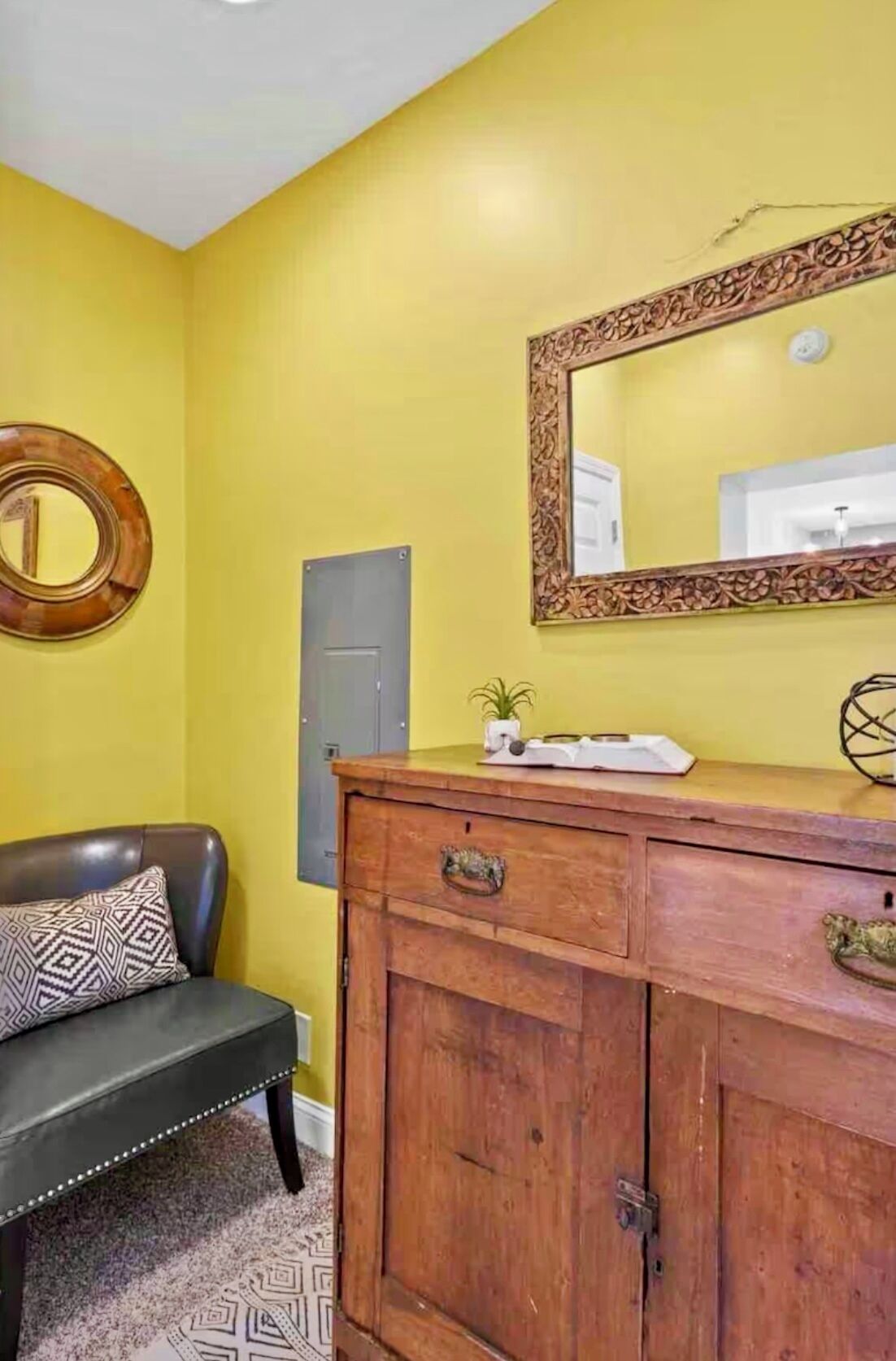A room with a yellow wall, a wooden cabinet with drawers, a mirror with an ornate frame, a black leather chair with a patterned pillow, and a small plant on the cabinet.