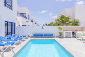 A pool area with blue lounge chairs, a small pool, and white buildings with blue accents. - Villa Elizabeth Rubicon (Yaiza)