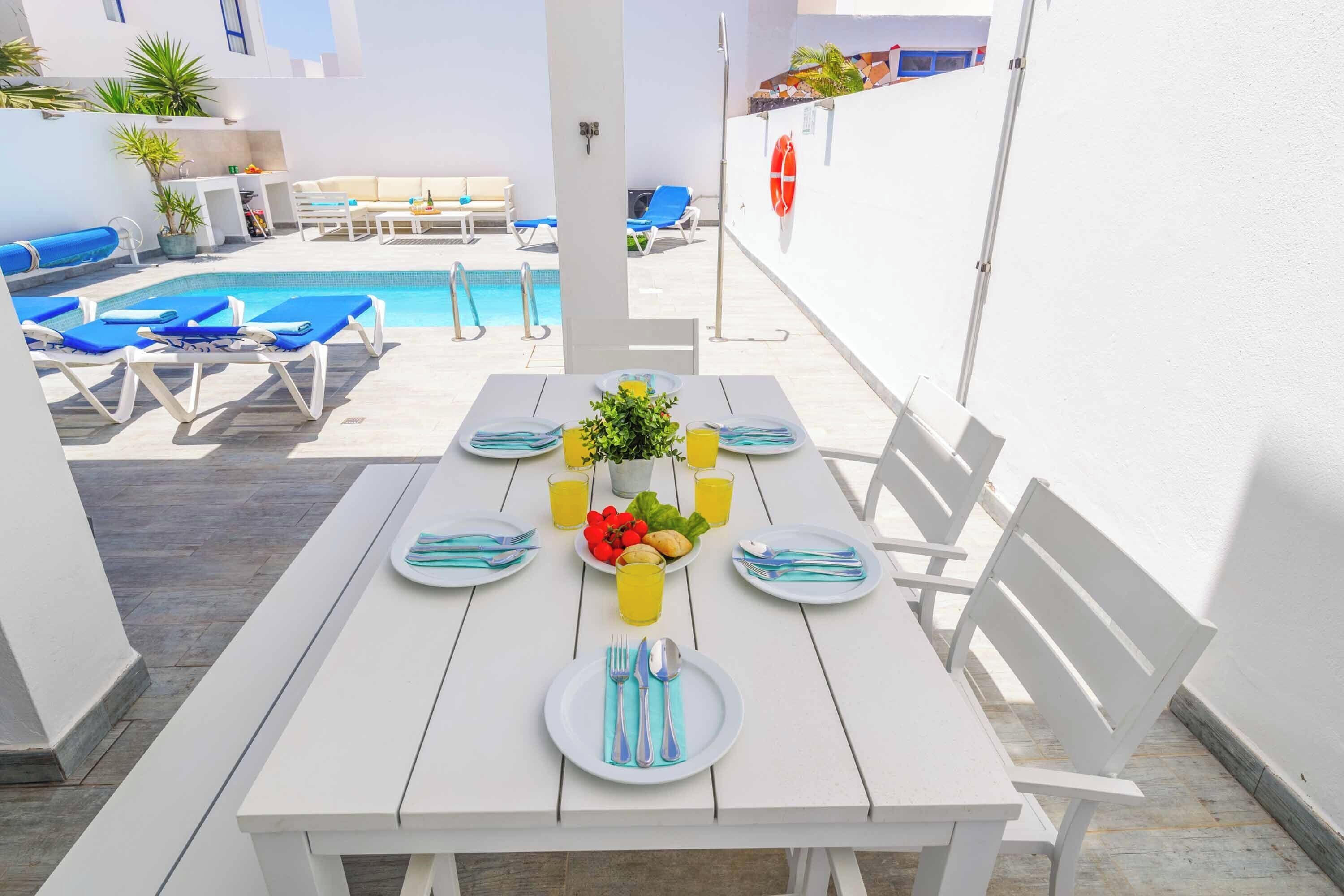 A white table set for four with plates, cutlery, and a fruit bowl, surrounded by white benches and a pool area in the background.
