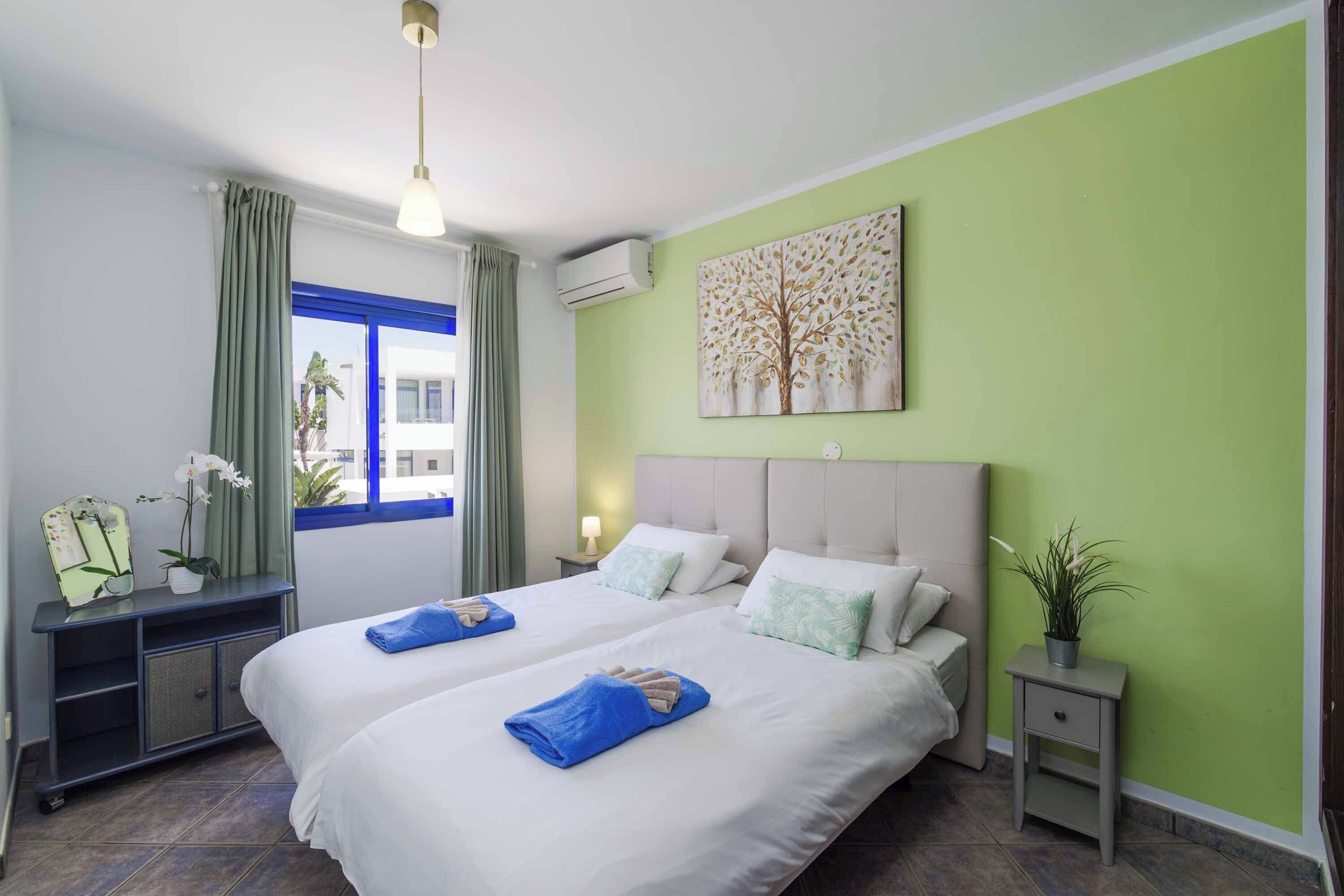 A hotel room with two beds, a desk, a nightstand, a window with curtains, an air conditioner, and a wall painting.