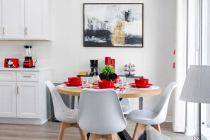 A modern dining area with a round table, white chairs, and red tableware. A coffee maker and a plant are on the table.