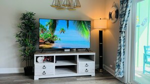 A living room with a flat-screen TV on a wooden TV stand, a potted plant, a lamp, and a view of the outdoors through a window.