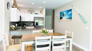 A modern kitchen with white cabinets, a wooden dining table, and chairs. There is a refrigerator, a coffee maker, and a small island with a sink.