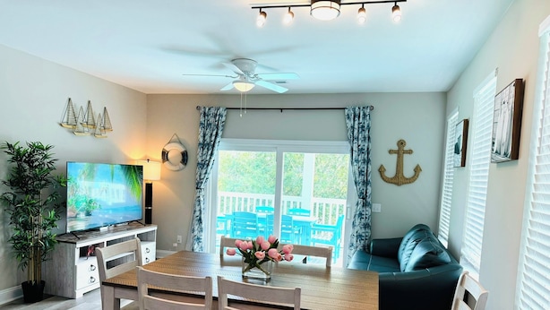A dining area with a wooden table and chairs, a television, and a view of a deck with blue chairs.