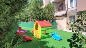 Zona de juegos infantiles con una casita y vehículos de juguete sobre césped artificial, rodeada de follaje y un edificio al fondo.